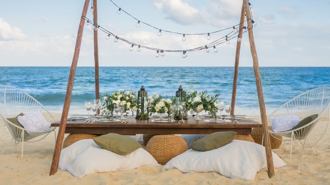 mesa con cena romántica para dos personas con flores y luces de colgantes en la playa con el mar en la parte posterior